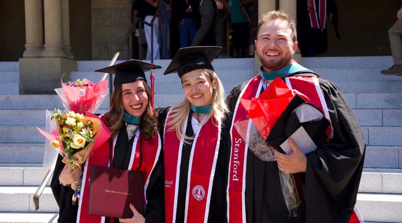 Tiffany Saade, Leticia Lie, and Kevin Klyman pose together in their graduation robes on the front steps of Encina Hall at Stanford University.