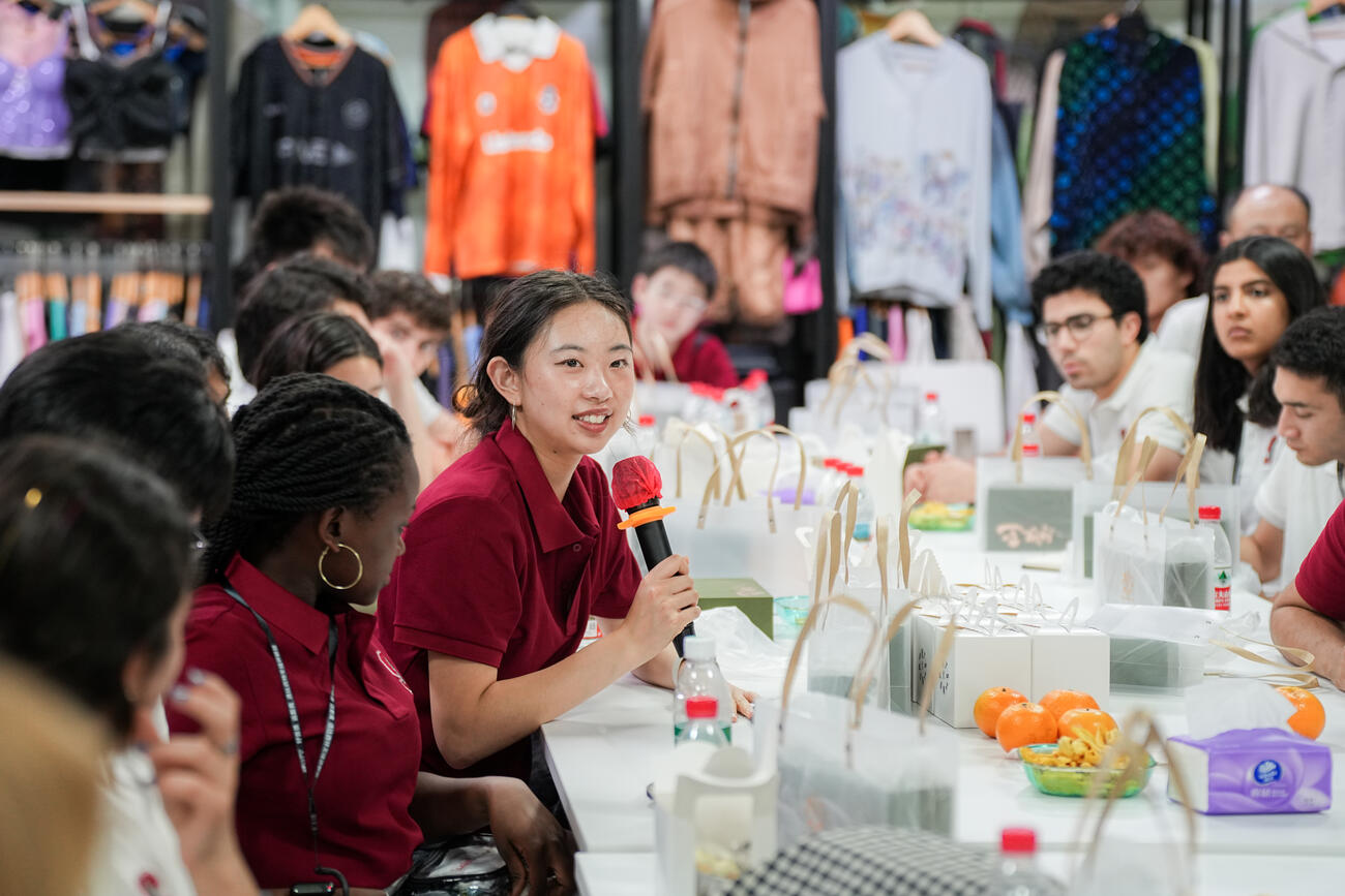 Student holds a microphone while seated at a table with other students in a garment factory.