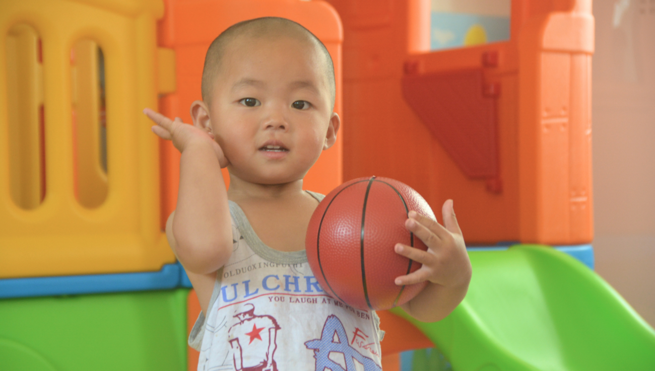 A toddler holds a small basketball in a play area.
