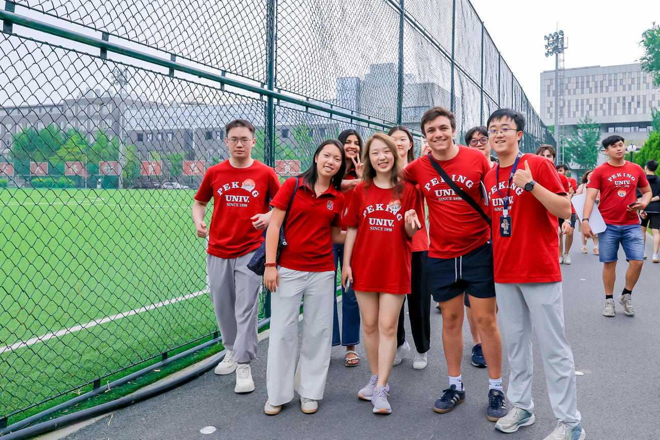 Students in Peking University shirts pose in front of a green sports field.