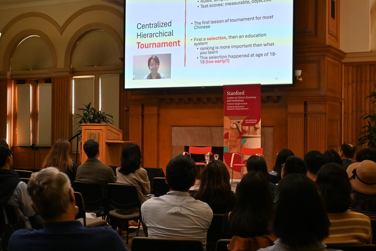 A full audience seated in front of a screen with a virtual speaker on it and presentation loaded.