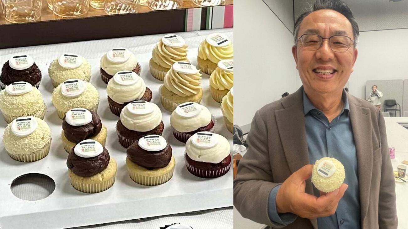 A tray of cupcakes decorated with the cover design of the book "The Four Talent Giants" and a photo of Gi-Wook Shin holding one of those cupcakes while similing to the camera.