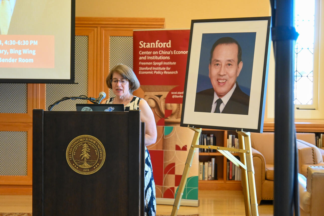 Julie Sweetkind-Singer speaks at the front of a room behind a podium and a large portrait of a man.