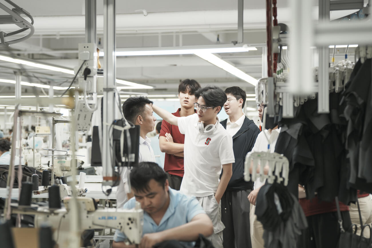 Students participate in a tour on the Yaying factory tour. A man sews in the forefront on a sewing machine.