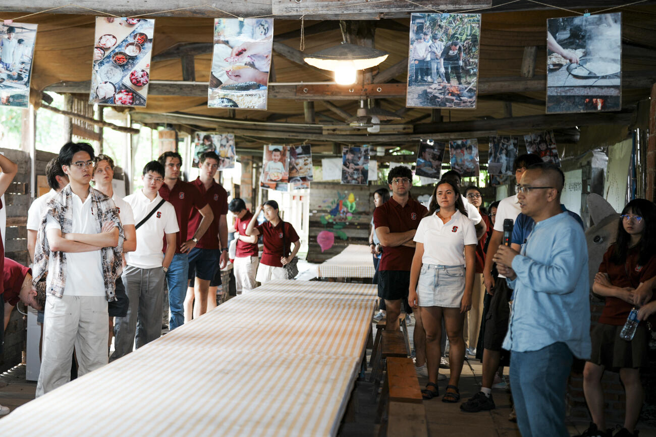Students stand in a crowded room with someone speaking on a microphone.