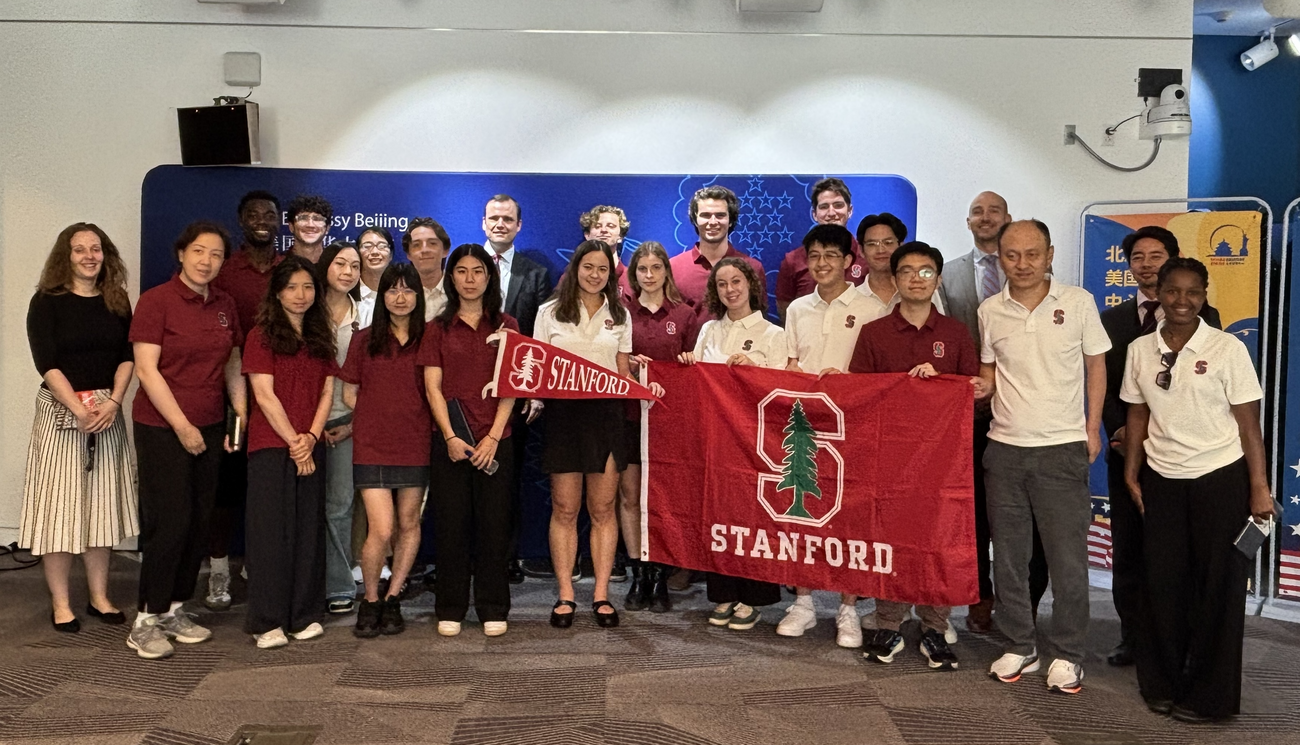 A group photo of students an faculty standing with a Stanford flag and an embassy banner.