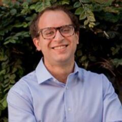 Eli stands in front of green bushes with his arms crossed and a slight smile on his face.