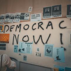 Wall plastered with posters and graffiti text reading "DEMOCRACY NOW"