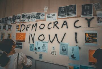 Wall plastered with posters and graffiti text reading "DEMOCRACY NOW"