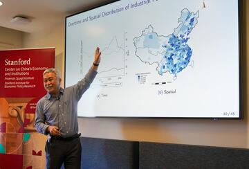 Hanming Fang presents in front of slides in a conference room.