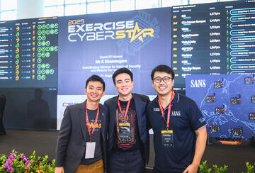 Photo of MIP Student RJ Teoh posing with two others in front of a banner that reads 2025 Exercise Cyber Star