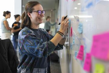 smiling woman standing next to a whiteboard filled with Post-Its