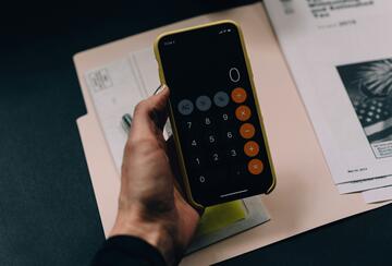 A hand holding an iPhone in a yellow case displaying the iOS Calculator app with a black screen, orange operator buttons, and gray function buttons, resting over papers and documents on a dark desk.