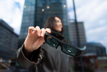 Close-up of smart glasses showing augmented reality interface while held by a woman in a modern urban environment.