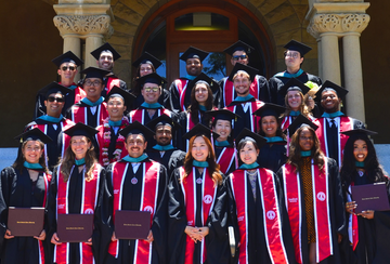 Graduates of the Ford Dorsey Master's in International Policy Class of 2026 pose on the steps of Encina Hall at Stanford University in their caps and gowns and diplomas.
