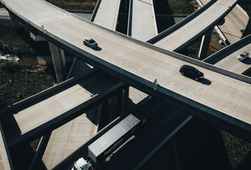 Aerial view of a multi-level highway interchange with sweeping curved overpasses crossing above one another. A few cars and a white semi-truck travel on the mostly empty concrete roads, while patches of vegetation and water are visible below the elevated ramps.