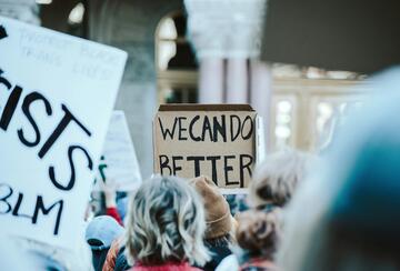 A crowd of people gathers at a protest, holding handmade signs. A cardboard sign in the center reads “WE CAN DO BETTER.”