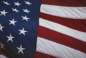 Close-up photo of a rippling American flag, with embroidered white stars on a blue field on the left and red-and-white stripes waving on the right.