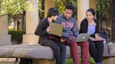 Three students conversing while sitting on a bench, two of them holding open laptops.