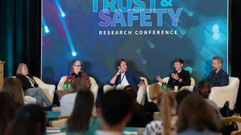 six people sit in white chairs on a raised stage in front of a turquoise and magenta background