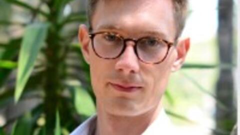 Headshot of Johannes Junge Ruhland. Ruhland is wearing glasses and a collared shirt in front of greenery.