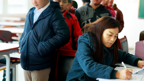 A teacher grades papers with a line of students behind her.