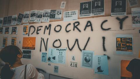 Wall plastered with posters and graffiti text reading "DEMOCRACY NOW"