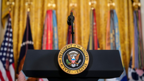 An empty lectern beairng the official seal of the United States in front of a row of flags.