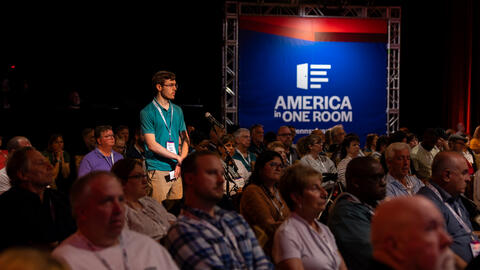 A participant stands at a microphone to speak during a large forum, while an audience listens in a conference setting with a backdrop reading “America in One Room.”