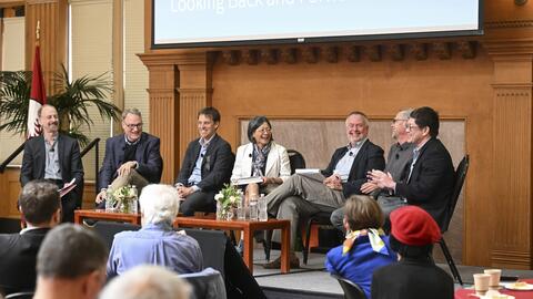 A panel of six scholars seated on stage in a conference room while in discussion in front of audience members.