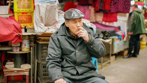 A man sits on a corner in China smoking with merchandise towering behind him.