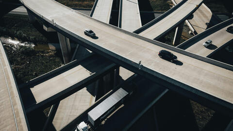 Aerial view of a multi-level highway interchange with sweeping curved overpasses crossing above one another. A few cars and a white semi-truck travel on the mostly empty concrete roads, while patches of vegetation and water are visible below the elevated ramps.