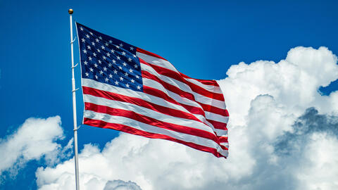 An American flag flying from a white pole against a bright blue sky with large white clouds.