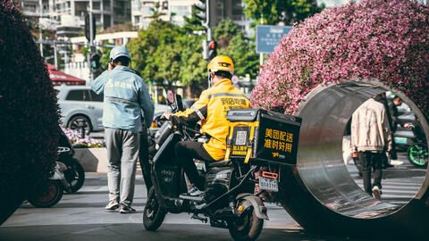 Two delivery workers, one on a scooter, wait outside a building in an urban setting in Shenzhen, China.