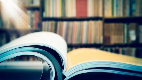 A beam of light shining on a stack of open journals set on a desk against the backdrop of shelves filled with books.