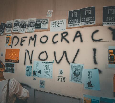 Wall plastered with posters and graffiti text reading "DEMOCRACY NOW"
