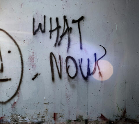 Graffiti on a concrete wall with a "meh" face and the words WHAT NOW?