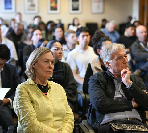 Audience members sit attentively during a lecture or seminar, with several people in the foreground listening closely while others take notes in a crowded room.