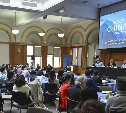 A room full of people seated at round tables facing a stage with five panelists seated behind a long table.