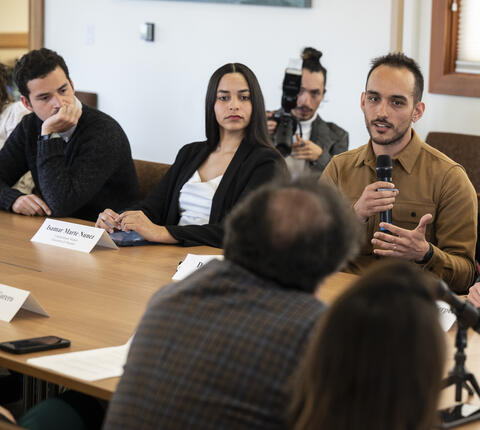 A man speaks into a microphone at a seated panel discussion with name placards and a small Colombian flag on the table, as other participants listen and take notes.