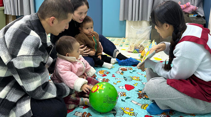 Parents hold two babies while caretaker reads to them.