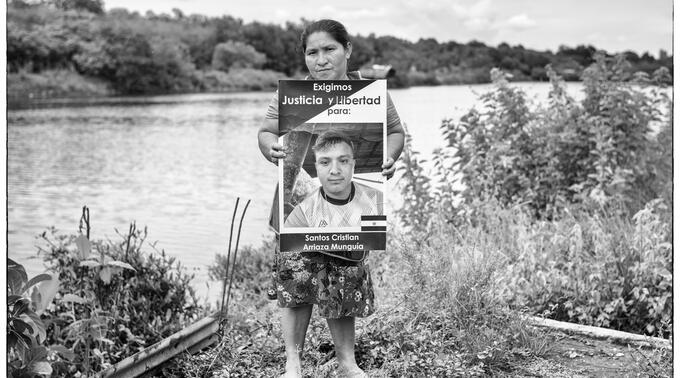 Black-and-white photo of a woman standing by a riverbank holding a sign reading ‘Exigimos Justicia y Libertad para: Santos Cristian Arriaza Munguía’ with his photo.