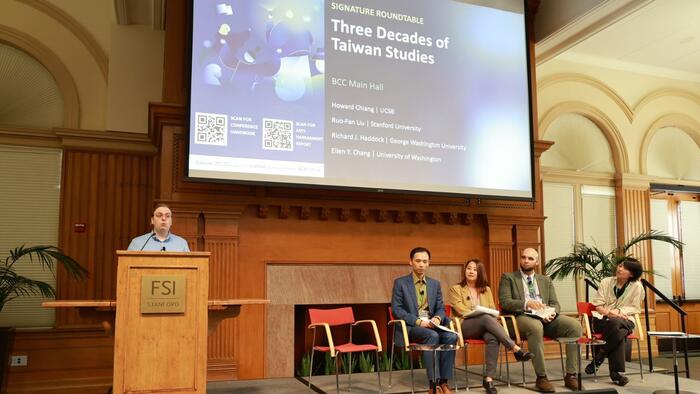 A presenter at a lectern and four panelists on stage in a conference room.