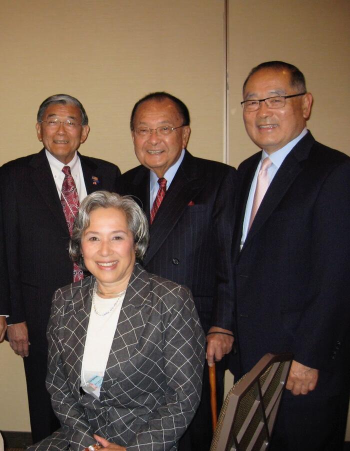 three people in suits standing in the back and one person sitting
