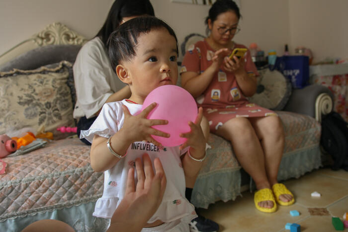 A toddler plays with a pink ball while their caregivers sit on the couch behind him looking at their phones.