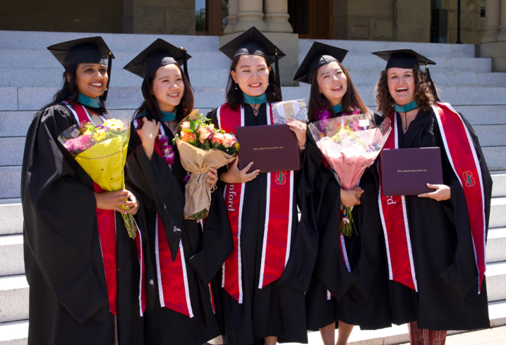 Sakeena Razick, Leticia Lie, Euysun Hwang, and Julie Tamura pose in their graduation caps and gowns on the steps of Encina Hall at Stanford University.