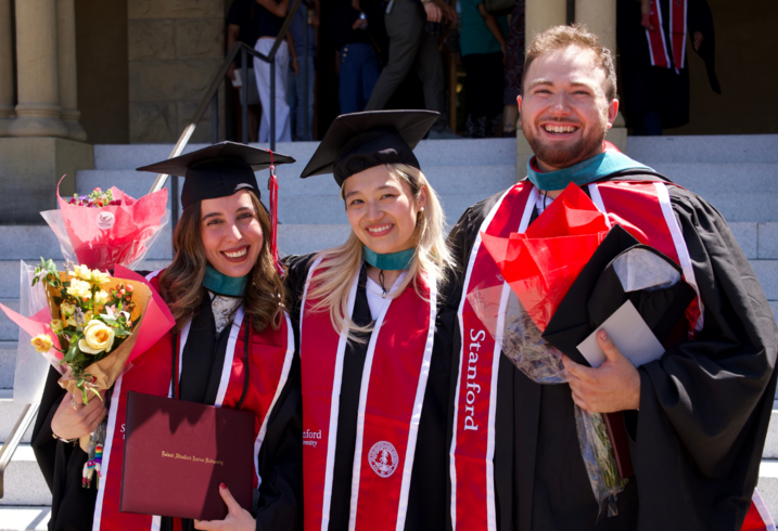 Tiffany Saade, Leticia Lie, and Kevin Klyman pose together in their graduation robes on the front steps of Encina Hall at Stanford University.