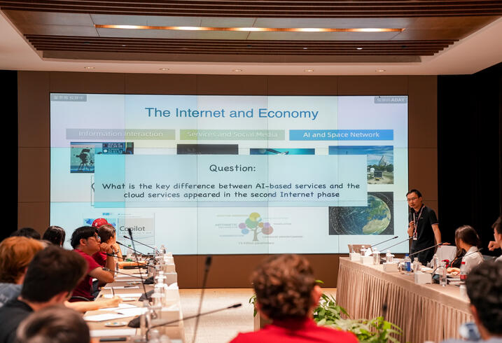 Students sit at a conference table looking at a digital screen that shows a presentation on AI and the economy.