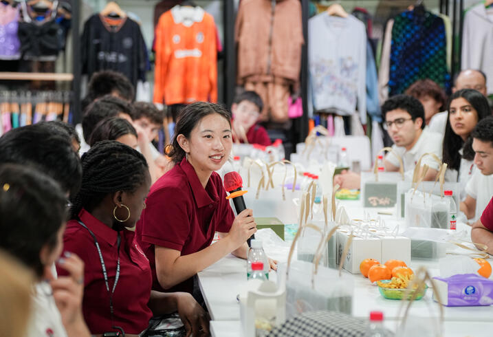 Student holds a microphone while seated at a table with other students in a garment factory.