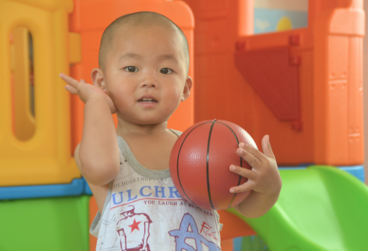 A toddler holds a small basketball in a play area.